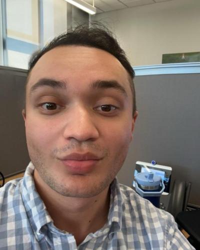 A man wearing a blue and white plaid shirt smiles at the camera from an office cubicle.