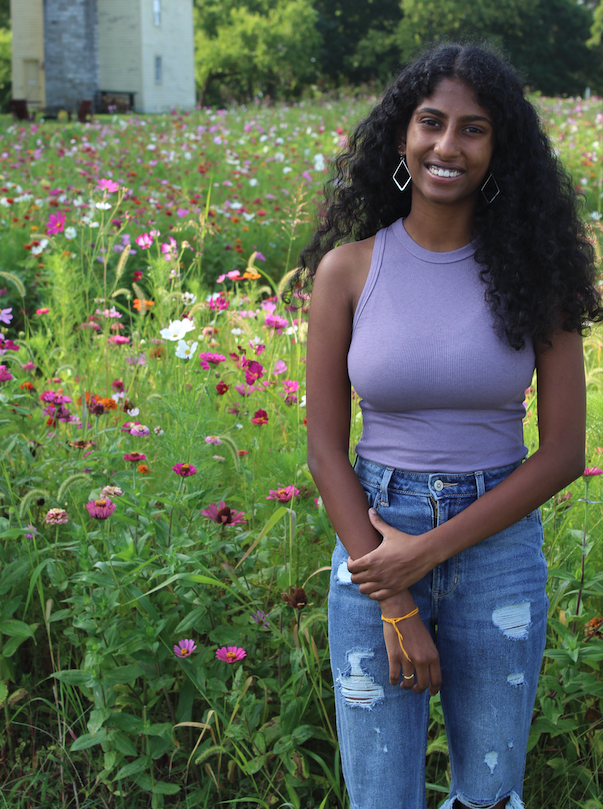 Diksha Satish stands smiling in a field of wildflowers, wearing a lavender sleeveless top and ripped jeans. A farmhouse is visible in the background under a sunny sky.